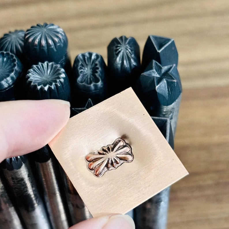 May include: A selection of metal stamps with diverse decorative patterns. A small, square copper-coloured metal piece is held up, showing an embossed floral design. The stamps are dark grey and feature various shapes, including stars and flowers.