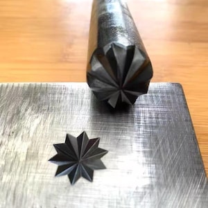 May include: A cylindrical metal stamping tool with a star-shaped tip featuring twelve points. The tool is positioned above a flat, brushed metal plate that displays a crisp, indented star impression matching the design of the tool's tip.