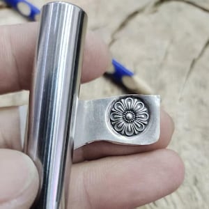 Puede incluir: Un tapón de botella de metal plateado con un mango cilíndrico y un diseño floral decorativo. El diseño floral está grabado en relieve en una placa rectangular unida al mango. El tapón está diseñado para encajar en la abertura de una botella.