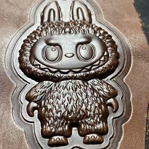 May include: A brown leather embossed design of a cartoon monster with large eyes, a wide grin, and furry body. The design is framed by a decorative border and is likely for crafting or leatherwork.