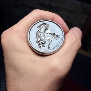 May include: A silver-colored, circular object with a raised relief of a rearing horse. The horse is detailed with flowing mane and tail. The object is held in a hand, and the background is dark.