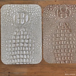 Puede incluir: Dos superficies rectangulares texturizadas con esquinas redondeadas. Una es plateada, la otra marrón. Ambas tienen un patrón de piel de cocodrilo. La superficie plateada está a la izquierda y la marrón a la derecha. El fondo es una superficie de madera.