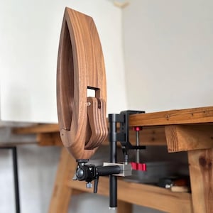 K&ouml;nnte beinhalten: Ein h&ouml;lzerner Holzbearbeitungsschraubstock mit einem einzigartigen, geschwungenen Design. Der Schraubstock besteht aus einem warm get&ouml;nten Holz und ist an einer schwarzen Metallklemme befestigt. Die Klemme ist auf einer h&ouml;lzernen Werkbank montiert.