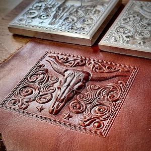 Puede incluir: Primer plano de un proyecto de artesanía en cuero con un diseño en relieve de una calavera de toro, rodeada de volutas florales y estrellas. Dos sellos metálicos con diseños similares se ven encima del cuero. El cuero es de color marrón intenso.