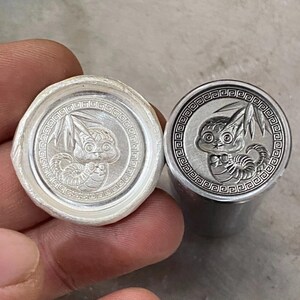 Könnte beinhalten: Zwei silberfarbene Wachssiegelstempel. Jeder Stempel zeigt ein detailliertes Relief eines stilisierten Wesens mit großen Augen und einem eingerollten Schwanz, umgeben von einem dekorativen Rand. Ein Stempel ist in einem Wachssiegel, der andere ist ein Stempel.
