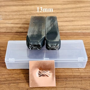 May include: Two metal stamps with a floral design, 13mm, next to a clear plastic case and a copper square with an embossed floral pattern. The stamps are dark grey and rectangular. The copper square has a raised floral design.