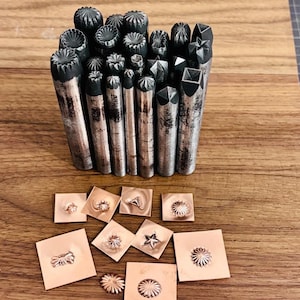 May include: A selection of metal stamping tools with diverse decorative patterns. The tools have cylindrical bodies and textured ends. Several stamped copper squares with embossed designs are also visible.
