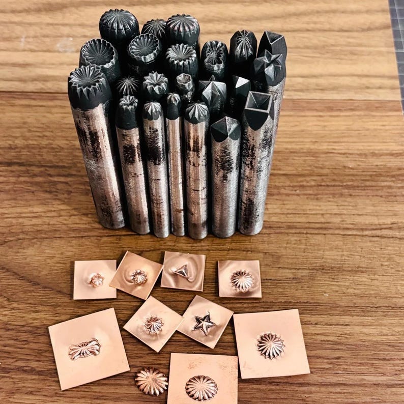 May include: A selection of metal stamping tools with diverse decorative patterns. The tools have cylindrical bodies and textured ends. Several stamped copper squares with embossed designs are also visible.