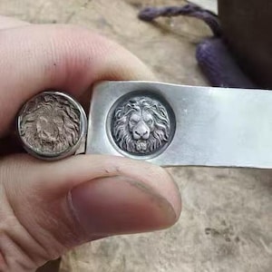 Może przedstawiać: Srebrzysty metalowy pierścień i pasujący prostokątny element metalowy, oba z wyraźnym, wypukłym reliefem głowy lwa z pełną grzywą. Wzór lwa jest wyśrodkowany na okrągłej powierzchni pierścienia oraz na prostokątnej metalowej płytce.
