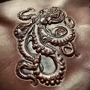 Peut inclure: Image en relief d'un poulpe sur une surface marron. Le poulpe est représenté avec des détails complexes, notamment ses tentacules, ses ventouses et sa peau texturée. L'image a un effet tridimensionnel.