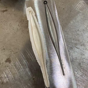 May include: Close-up of two elongated, teardrop-shaped molds. One is a light beige color, and the other is a dark gray. Both have a small circular hole at the top. The molds are set in a metal base.