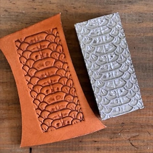 Puede incluir: Una pieza de cuero marrón con un patrón de escamas de cocodrilo estampado junto a un sello de metal plateado con el mismo diseño. El cuero es marrón cálido y el sello es rectangular.