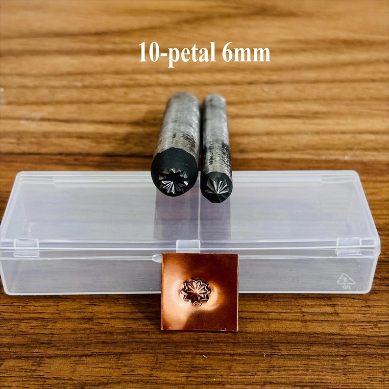 May include: Two metal stamping tools with 10-petal designs, 6mm in size, are displayed next to a clear plastic case and a stamped copper square. The tools are cylindrical with a dark, patterned end. The copper square shows the stamped floral design.