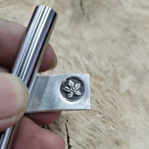 May include: A silver metal stamp with a maple leaf design. The stamp is circular and set on a rectangular piece of metal. A cylindrical metal tool is held nearby.