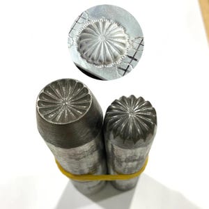 May include: Close-up of two metal stamping tools with floral designs. The tools are cylindrical with detailed, radial patterns on the stamping surfaces. A circular stamped impression is also visible, showcasing the design's final appearance.