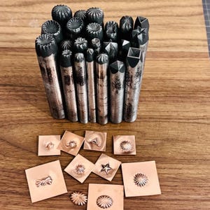 May include: A selection of metal stamping tools with diverse decorative patterns. The tools are cylindrical with textured ends. Several stamped copper squares with floral and geometric designs are also shown.