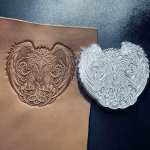 May include: A silver-colored metal eagle stamp featuring intricate feather and talon details, positioned next to a tan leather sheet that displays a deeply embossed, identical eagle design created by the stamp's impression.