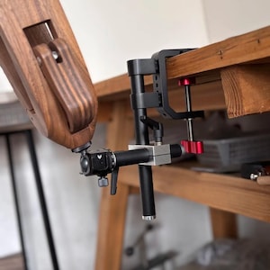 K&ouml;nnte beinhalten: Eine schwarze und silberne Kameraklemme mit rotem Einstellknopf ist an einer Holzoberfl&auml;che befestigt. Ein Holzarm ist mit der Klemme verbunden. Die Klemme dient zum Halten einer Kamera oder anderer Ger&auml;te.