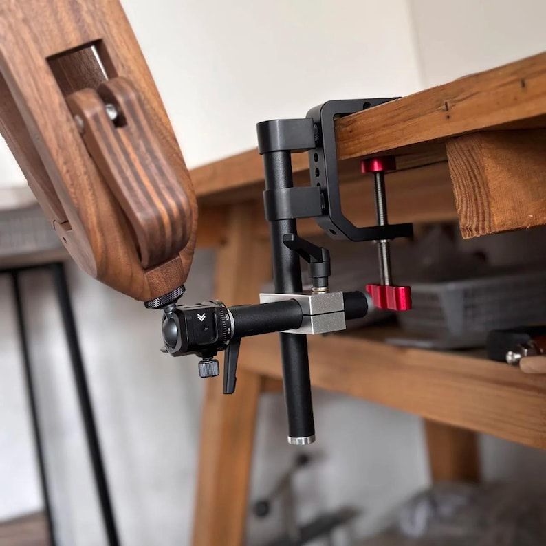 K&ouml;nnte beinhalten: Eine schwarze und silberne Kameraklemme mit rotem Einstellknopf ist an einer Holzoberfl&auml;che befestigt. Ein Holzarm ist mit der Klemme verbunden. Die Klemme dient zum Halten einer Kamera oder anderer Ger&auml;te.