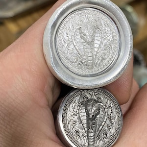 Könnte beinhalten: Zwei silberfarbene Metallobjekte mit einem detaillierten Kobra-Design. Die Kobra ist auf einer kreisförmigen Basis zentriert, umgeben von kunstvollen Mustern. Das obere Objekt hat einen breiteren, abgerundeten Rand, das untere ist kompakter.