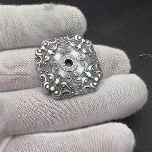 May include: A square, silver-colored pendant with an intricate, swirling design. The pendant features a central circular element with a geometric pattern and a hole in the center. The edges of the pendant are rounded, and the overall design is ornate.