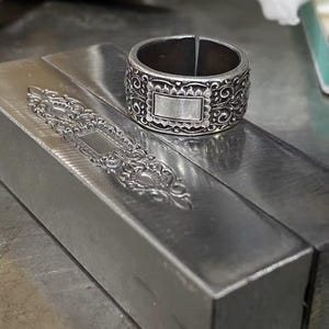 May include: A silver-colored ring with an ornate design featuring a rectangular center and scrollwork. The ring is wide and has a split in the band. The ring is displayed on a metal surface.