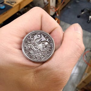 May include: A silver-colored coin featuring an octopus design. The coin has intricate details of the octopus and ocean waves. The coin is round and has a detailed border.