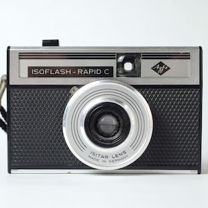 May include: A silver and black Agfa Isoflash-Rapid C camera with a silver Isitar lens. The camera has a black textured body and a silver top plate with the Agfa logo and the text "Isoflash - Rapid C".