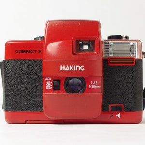 Peut inclure: Un appareil photo argentique 35 mm Hakimg Compact E rouge avec une poignée en simili cuir noir et un flash intégré. L'appareil photo possède un objectif avec une ouverture de 1:3.5 f=38 mm et un cadran de réglage ASA.