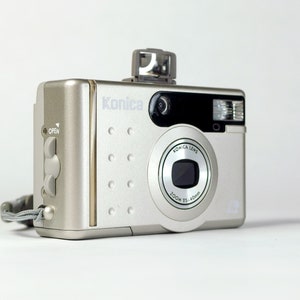 May include: Silver Konica digital camera with a pop-up flash. The camera has a zoom lens and the text "Konica Lens Zoom 35-40mm". The camera has a wrist strap attached and the word "Open" is visible.