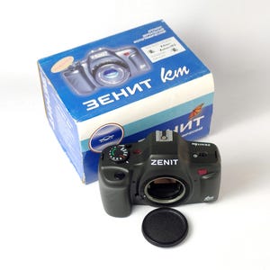 May include: A black Zenit camera with a lens cap. The camera has a silver dial and a red button. The camera is sitting on a white surface. The camera is in its original box, which is blue and white with the word "Zenit" in large letters.