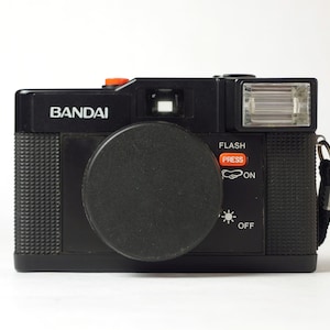 Peut inclure: Un appareil photo en plastique noir avec un capuchon d'objectif noir et un flash. L'appareil photo porte l'inscription "BANDAI" sur le devant. Le flash a un bouton rouge qui indique "FLASH PRESS" et un interrupteur qui indique "ON" et "OFF".
