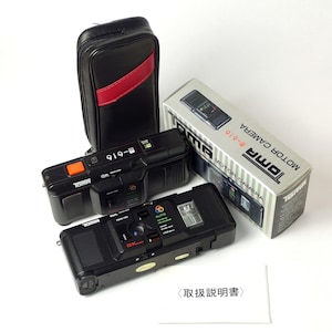 Puede incluir: Dos cámaras de película negras con una funda de transporte negra y una caja blanca con el texto "TOMA MOTOR CAMERA 8-616" impreso en ella. Las cámaras están etiquetadas como "TOMA" y tienen un botón rojo en el lateral.