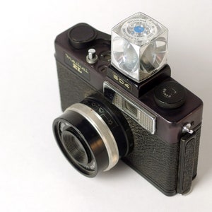 Peut inclure: Un appareil photo argentique noir et argenté 304 avec un flash. Le flash est un flash Sylvania Blue Dot en forme de cube. L'appareil photo a un objectif avec une bague argentée et un capuchon d'objectif noir.