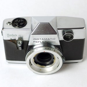 May include: A silver and black Kodak Instamatic Reflex camera with a lens ring showing aperture settings from 22 to 8. The camera has a black leatherette covering and a silver top plate with the words "Instamatic Reflex Camera" printed on it.