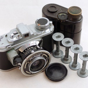 Op de afbeelding: Een zilveren en zwarte 35mm filmcamera met een zwarte lensdop en een zwarte filmhouder. De camera heeft een zilveren body met een zwarte lederlook bekleding. De lens is gemarkeerd met "Photovit" en heeft een zilveren ring met markeringen voor diafragma en sluitertijd. De camera heeft ook een zilveren filmtransportknop en een zilveren ontspanknop.
