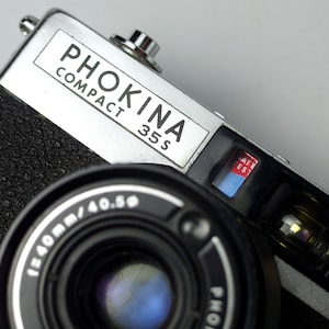 以下が含まれることがあります： 上部に「Phokina Compact 35S」の文字が書かれた、銀色と黒色の35mmフィルムカメラ。カメラは、銀色のリングが付いた黒いレンズと、黒い革張りカバーを備えています。