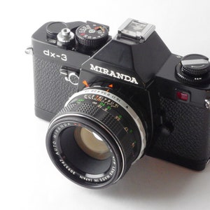 May include: A black Miranda dx-3 35mm film camera with a Miranda Auto 50mm f/1.7 lens attached. The camera has a silver hot shoe and a black leatherette covering.