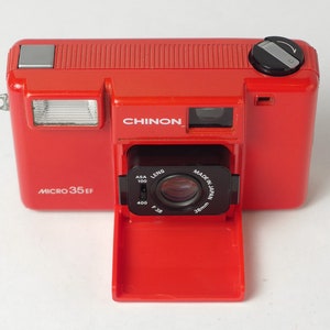 Peut inclure: Un appareil photo argentique rouge Chinon Micro 35EF avec un objectif noir et un flash. L'appareil photo est fabriqué au Japon et possède un objectif de 38 mm.