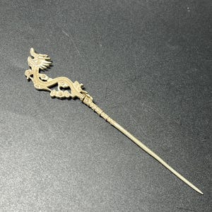 May include: A silver-toned metal hair pin featuring an intricate, sculpted dragon design at the top. The dragon figure is elongated with detailed scales and cloud-like motifs, transitioning into a long, slender, straight pointed metal shaft.
