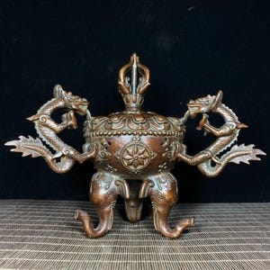 以下が含まれることがあります： ブロンズ色の金属製香炉。側面には2つの龍の形をした取っ手があり、台座には3つの象の頭の形をした脚、中央の蓋の上部には垂直な飾りが付いています。本体には円形の花のモチーフが装飾されています。