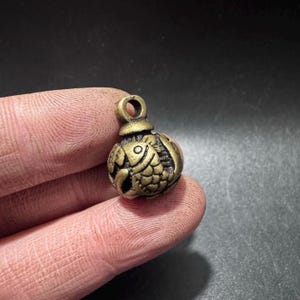 May include: A small, round, antique-toned brass charm featuring a detailed embossed fish design on its surface. The charm has a prominent circular loop at the top for attachment, with a textured, weathered metallic finish and visible scale patterns.