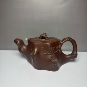May include: A dark brown clay teapot with a textured, organic shape resembling a tree trunk. The body features embossed floral branch motifs, a curved spout, and a handle with a gnarled texture. A matching lid with a small, sculpted knob sits on top.