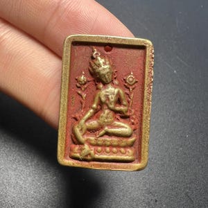 May include: A rectangular pendant featuring a gold-toned relief of a seated figure on a textured red background. The figure is adorned with a crown and flanked by two floral motifs. The pendant has a small hole at the top for suspension.