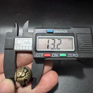 Puede incluir: Una peque&ntilde;a cuenta de metal de color lat&oacute;n, de forma redonda, con un dise&ntilde;o de pez en relieve. La cuenta est&aacute; sujeta por un calibrador digital negro que muestra una medida de 13,2 mm en su pantalla, indicando el di&aacute;metro del objeto.