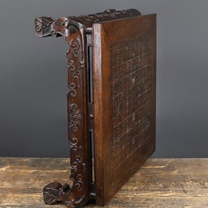 May include: A dark brown wooden board game table featuring an intricate grid pattern etched onto the flat surface. The side panels display elaborate carved scrollwork and decorative motifs, supported by curved legs with detailed relief carvings.
