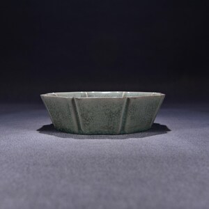 May include: A shallow, celadon-colored ceramic bowl with a lobed, flower-like rim and a distinct crackle glaze finish. The bowl features vertical indentations along its exterior walls, creating a faceted appearance with a muted green hue.