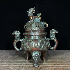 May include: A bronze-toned metal incense burner featuring a lid topped with a mythical creature. The body is adorned with embossed dragon motifs, two side handles shaped like dragon heads, and three legs with carved animalistic facial details.