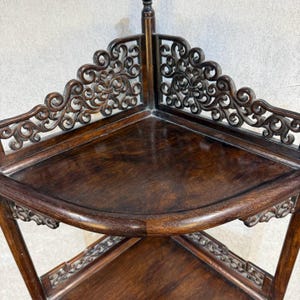 May include: A dark brown wooden corner shelf featuring two triangular tiers with smooth surfaces. The unit is accented by intricate, openwork scroll carvings along the back edges of each shelf, providing a decorative border for the corner structure.