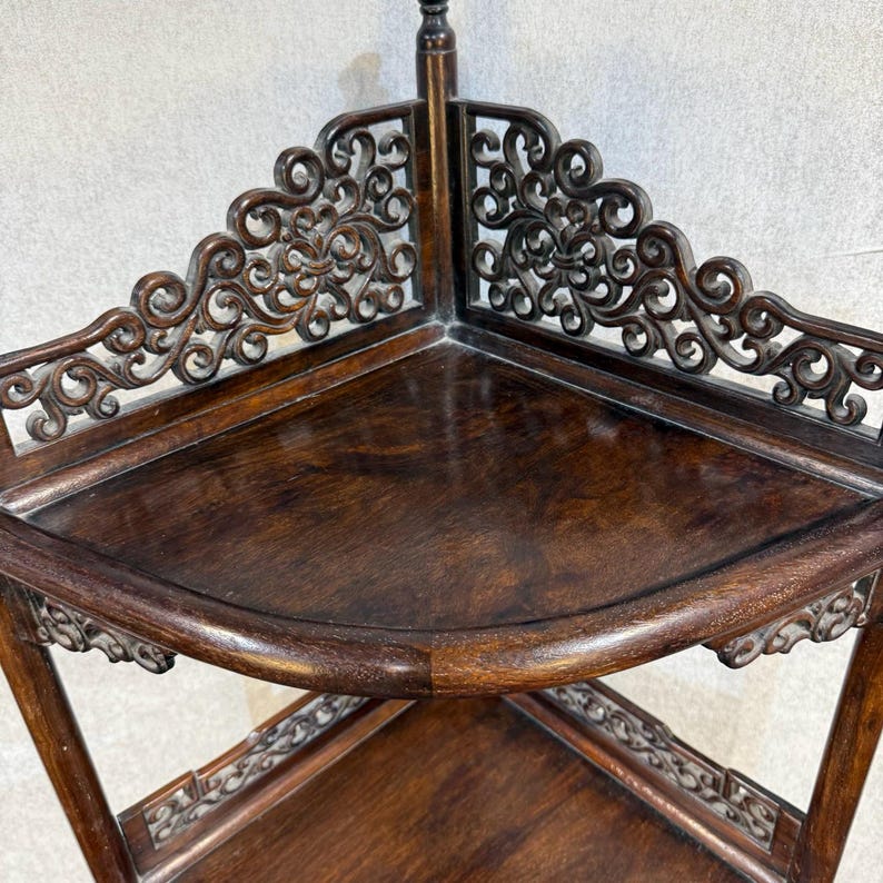 May include: A dark brown wooden corner shelf featuring two triangular tiers with smooth surfaces. The unit is accented by intricate, openwork scroll carvings along the back edges of each shelf, providing a decorative border for the corner structure.